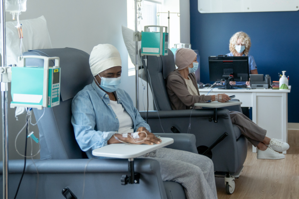 Two women sitting in seperate chairs recieving their cancer treatment. You can also see a nurse in the background on a computer.